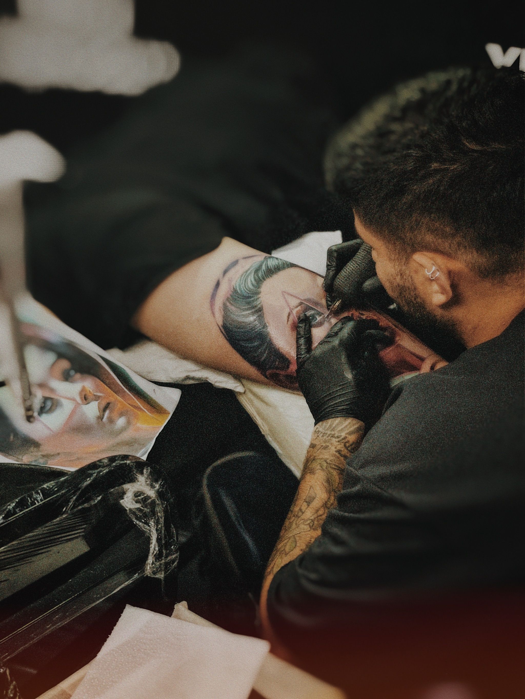 Tatuador aplicando retrato realista colorido com foto de referência posicionada ao lado